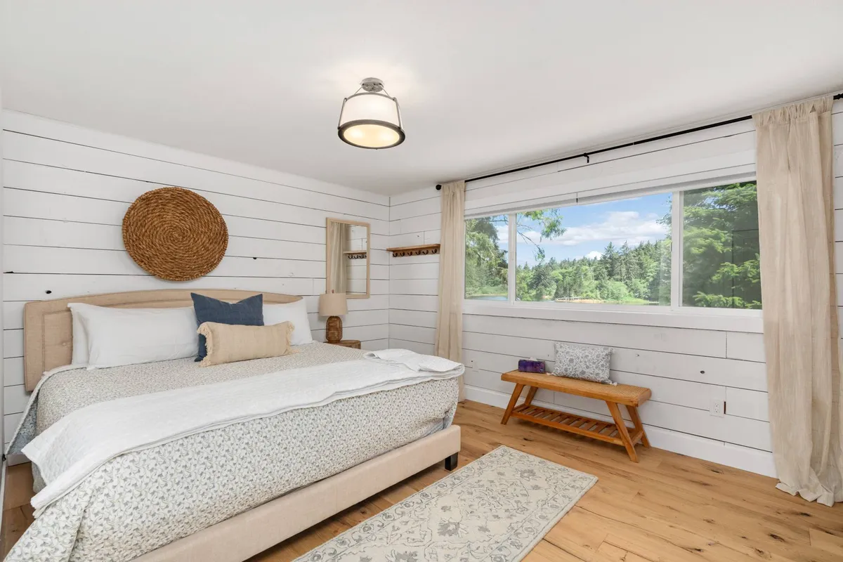 A cozy bedroom featuring a comfortable bed beside a window that opens to tranquil outdoor scenery. Woven wall decor and a simple nightstand add warmth, while the window view suggests a seamless connection to nature and a peaceful atmosphere for unwinding.