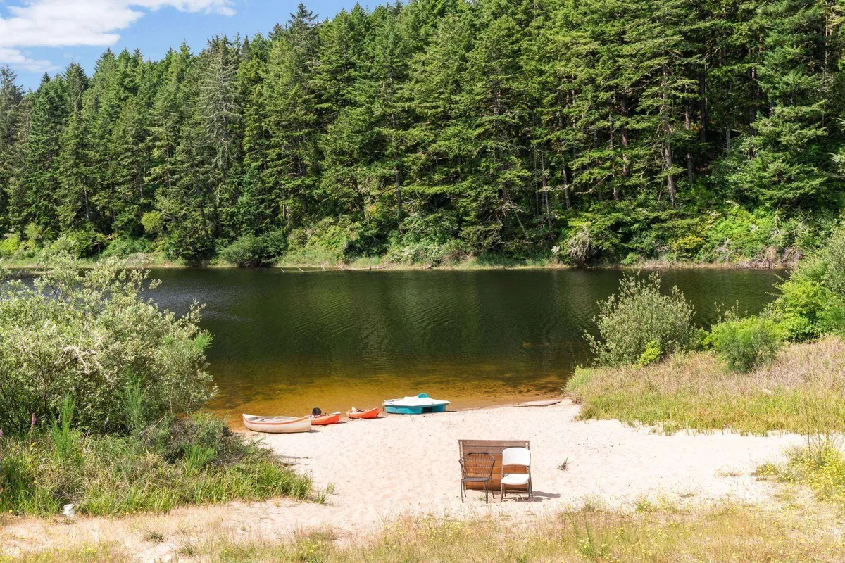 A peaceful lakeside scene featuring a calm lake bordered by lush greenery, with colorful kayaks lined up along the shore and a wooden chair inviting relaxation. The soft light suggests either early morning or evening, with gentle reflections on the water creating a serene atmosphere.