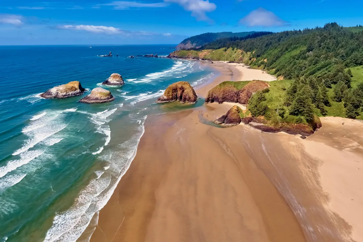 A tranquil beach scene showcasing iconic rock formations, gently rolling waves, a sandy beach, and green hills in the background. The natural beauty and serenity of the Oregon coast are on full display, inviting exploration and outdoor activity.