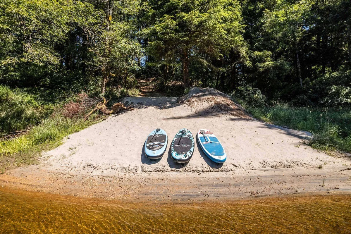 A sandy beach set along the lake's edge, with three paddleboards resting on the shore. The surrounding area features lush greenery, suggesting privacy and a natural setting ideal for outdoor activities like paddleboarding or swimming.