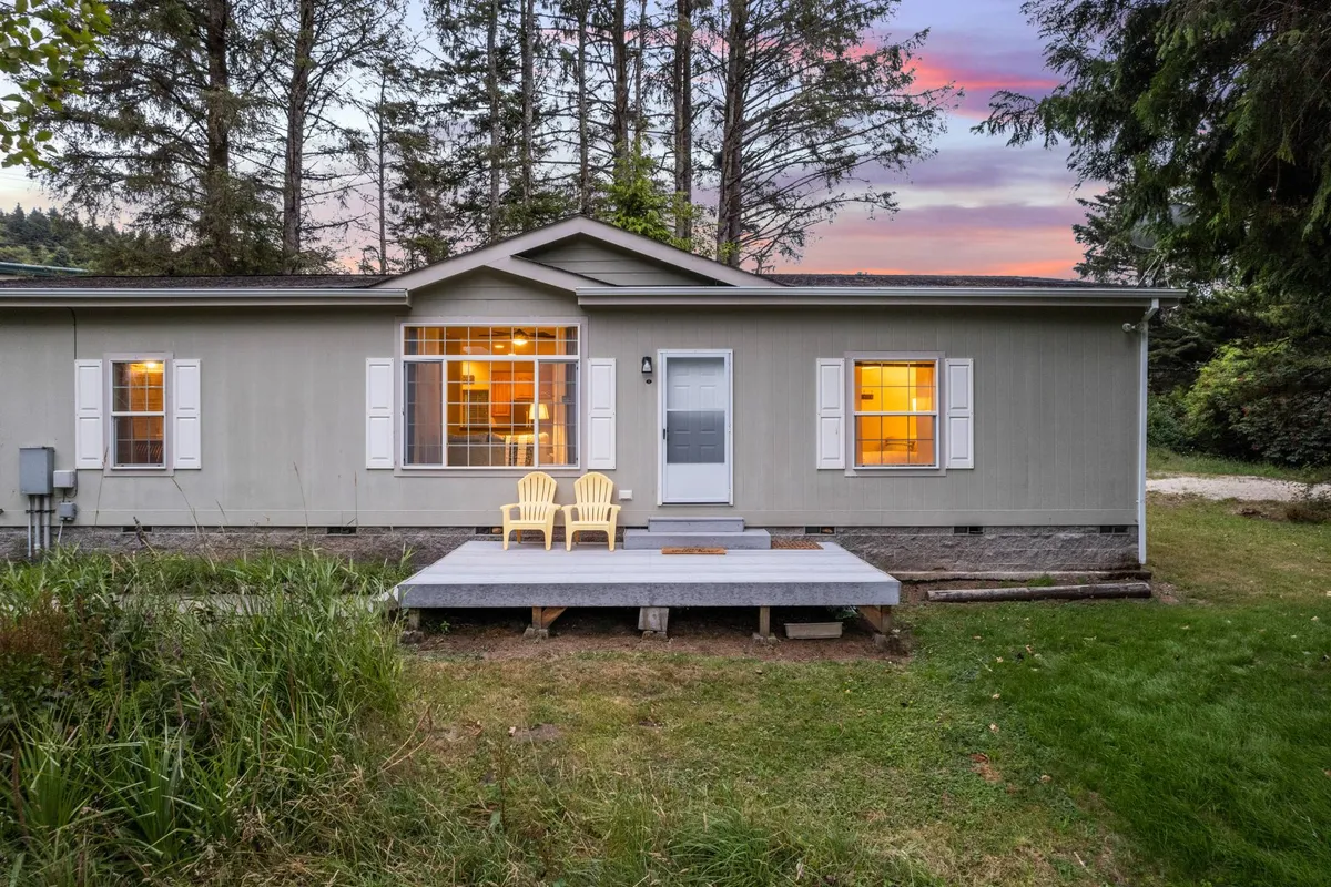 A charming exterior shot of one of the estate's homes, featuring a welcoming front porch with bright yellow chairs, large windows, and mature trees all around. The setting feels serene and inviting, ideal for enjoying nature or gathering with friends and family outdoors.