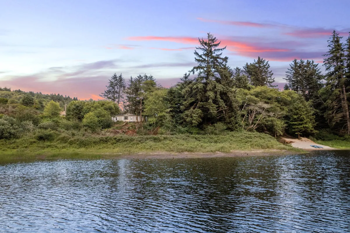 A serene view of the lake with the estate's house nestled among the trees. The tranquil water, sky, and abundant greenery evoke a sense of peace and natural beauty, while the property itself is clearly visible, reinforcing the 'private lake access' and expansive outdoor space.