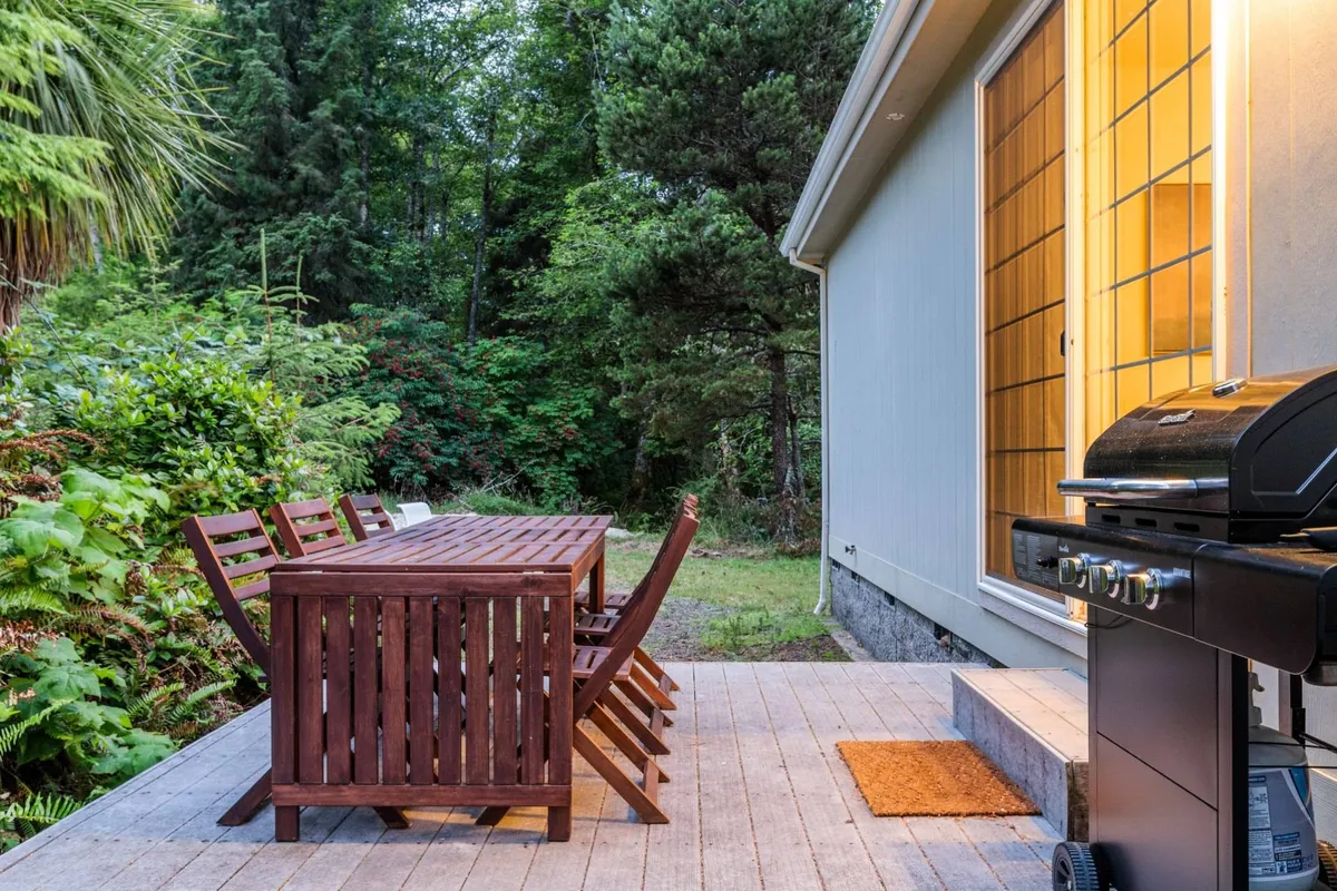 An inviting outdoor dining area with a large wooden table, BBQ grill, and lush greenery surrounding the space. Warm light emanates from the house, suggesting a cozy atmosphere for social gatherings, sunset dinners, or group celebrations.