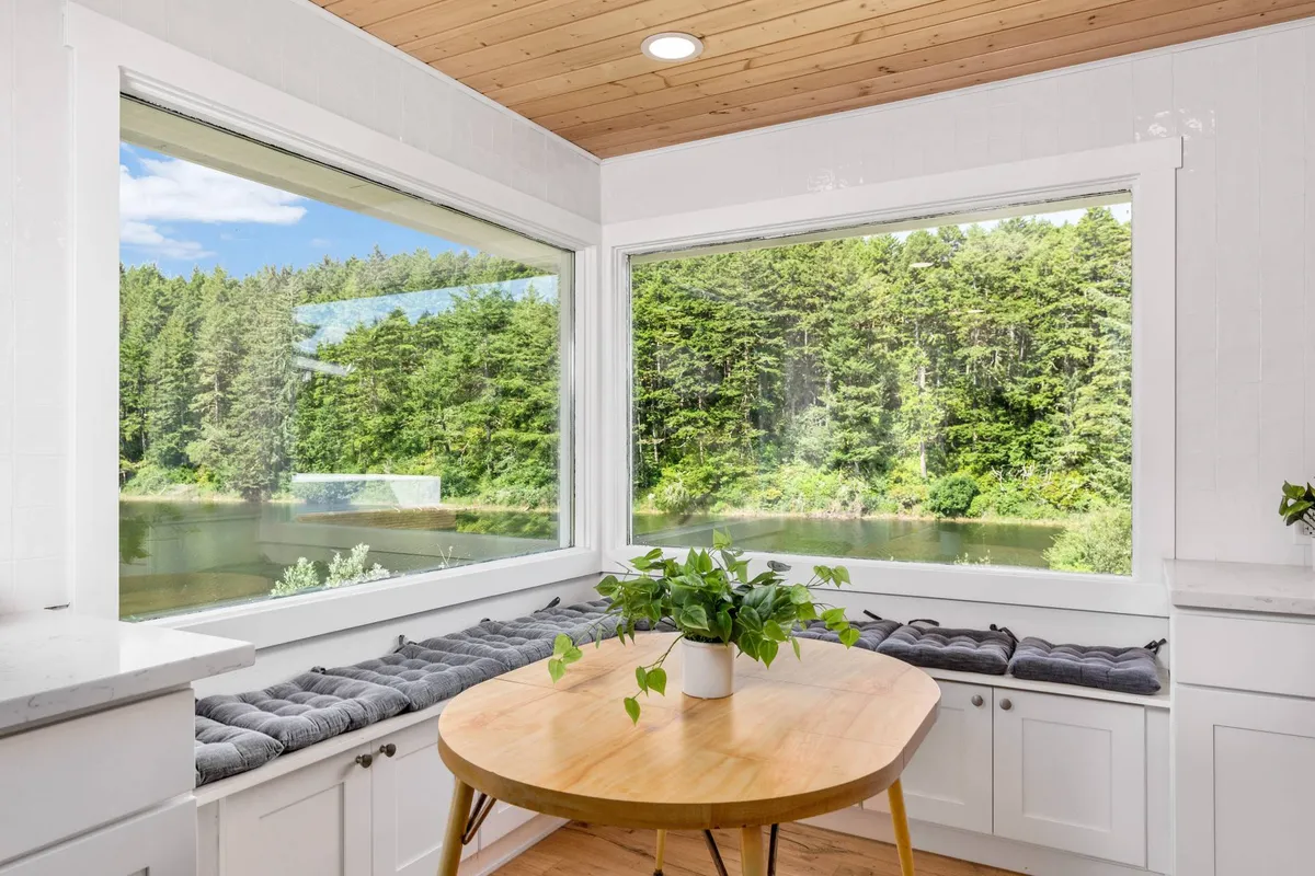 A peaceful dining area with a round wooden table and greenery in a vase is framed by expansive windows that offer sweeping views of the lake and forest outside. The soft natural light creates a serene atmosphere perfect for contemplation or enjoying a meal while surrounded by nature.