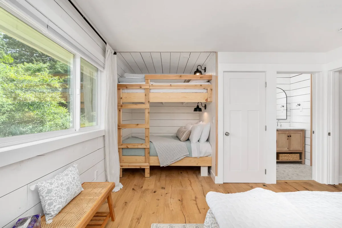 A cheerful bunk room featuring a bunk bed, a wooden bench, and a large window. Sunlight streams into the room, creating a space that is both fun for kids and functional for families. The design encourages relaxation and shared moments.