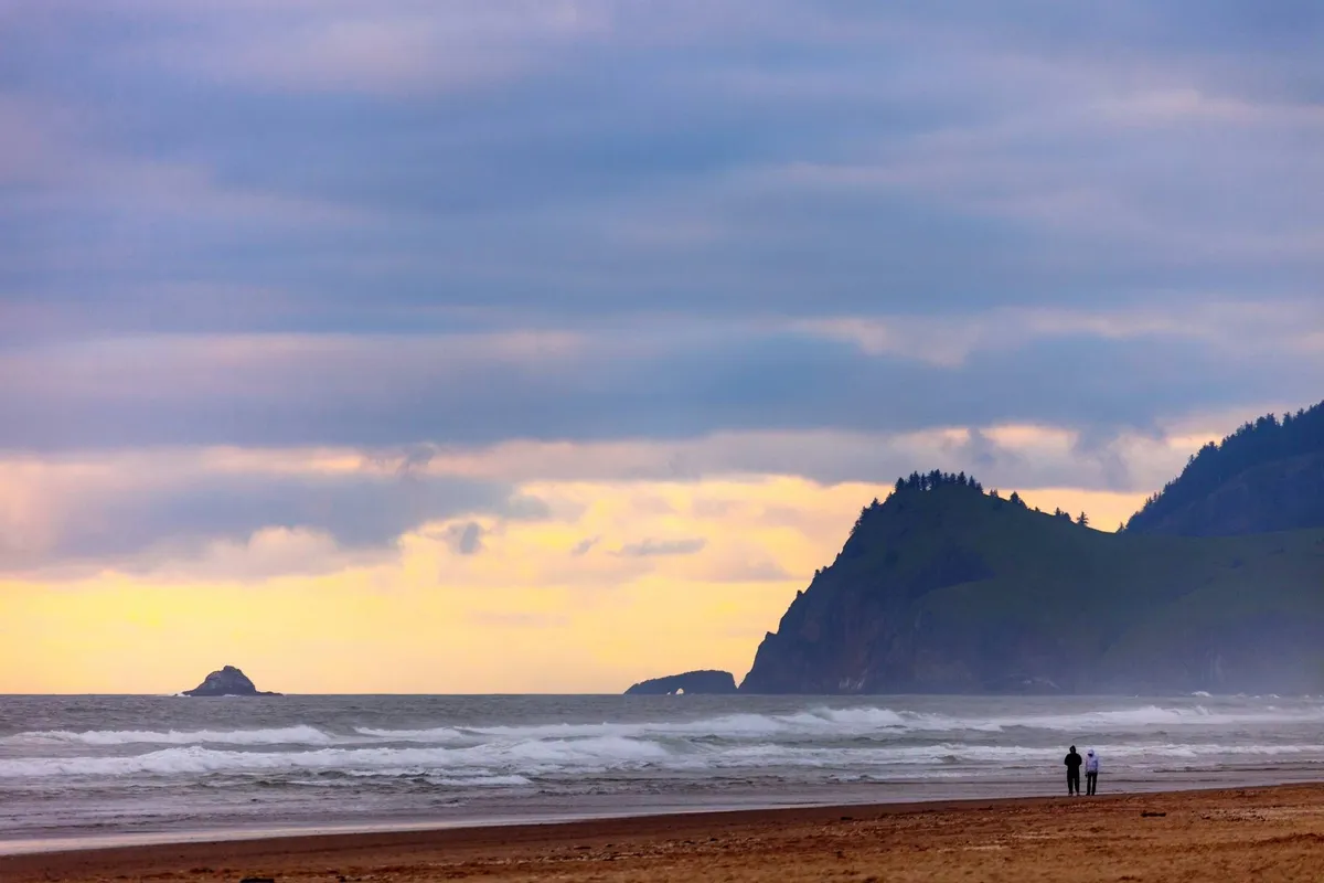An exterior view featuring gentle ocean waves, dramatic rock formations, and silhouetted figures walking along the shore at sunset. The reflective mood and natural setting evoke the estate's proximity to water and the peaceful, contemplative experiences available.