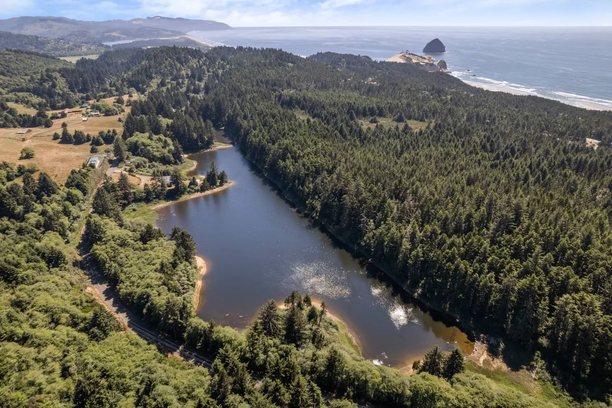The hero image showcases a tranquil private lake surrounded by lush forest, with the shimmering Pacific Ocean and iconic coastal rock formations visible in the distance. The sunset casts a warm glow over the water, evoking serenity and inviting outdoor activities such as fishing, hiking, and stargazing. Wildlife and natural beauty are abundant, emphasizing the secluded and exclusive nature of the estate.