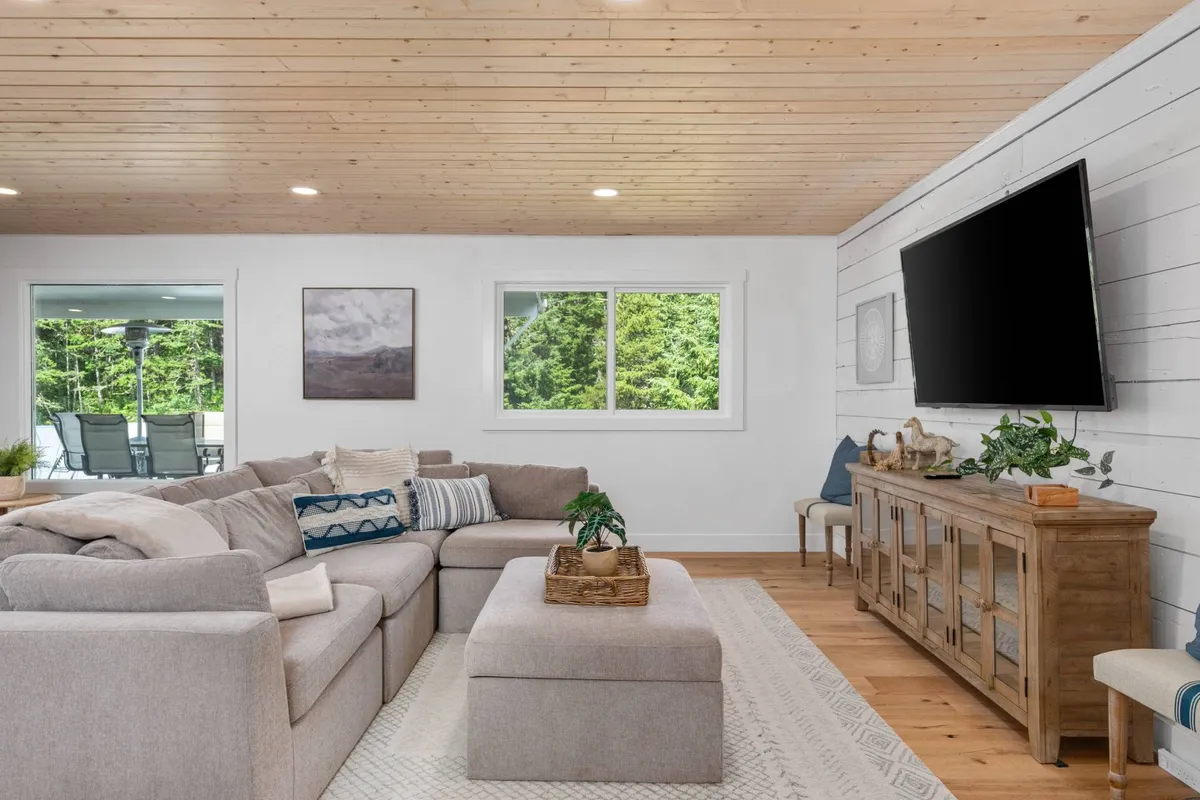 A large, comfortable sectional sofa is the heart of this living room, set around a wooden coffee table. Through the oversized window, lush greenery creates a calming backdrop, reinforcing the tranquil and welcoming mood. The room is ideal for relaxing with family, gathering with friends, or simply unwinding after a day outdoors.