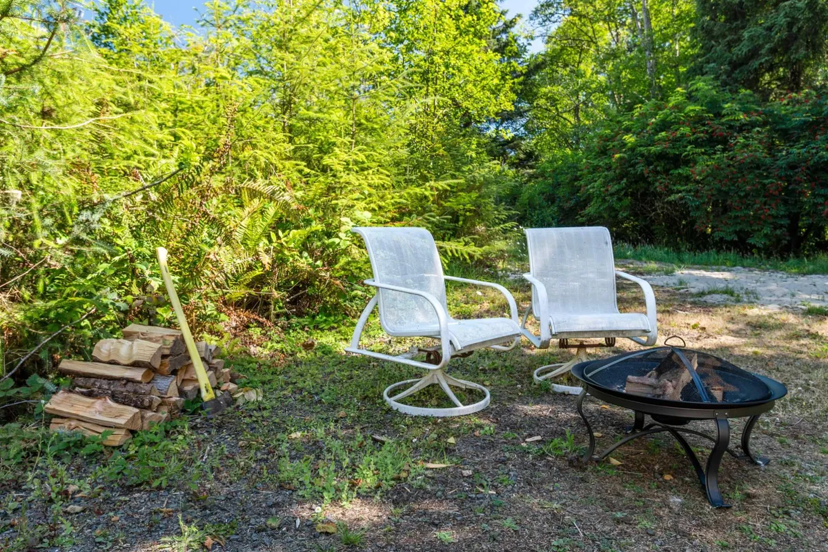 A cozy outdoor scene featuring two white rocking chairs set around a fire pit with a stack of firewood nearby. The lush greenery in the background and the tranquil setup suggest evenings spent socializing, stargazing, or sharing stories around the fire.