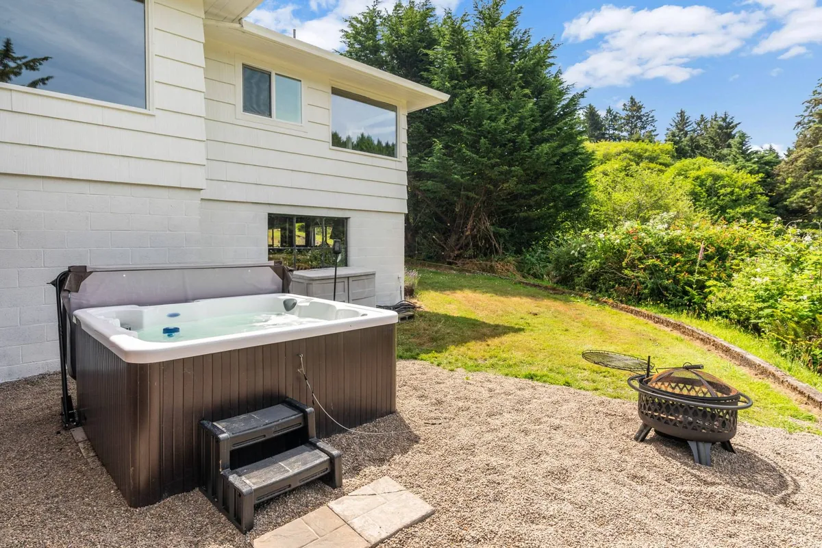 An inviting exterior setting featuring a bubbling hot tub and a fire pit surrounded by landscaped gardens, all set against a backdrop of natural beauty. The scene suggests relaxation, socializing, and evening gatherings under the stars.