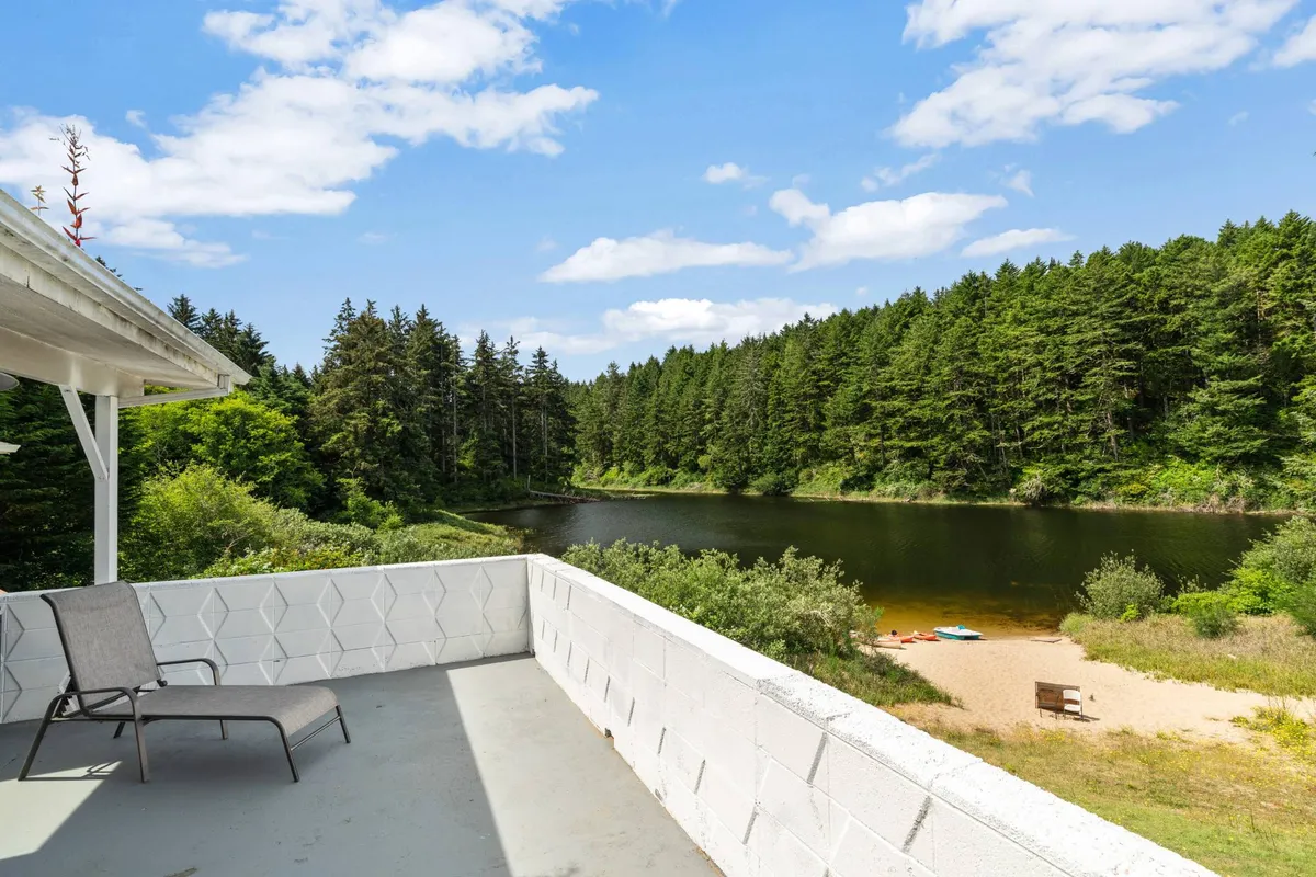 A broad view of the tranquil lake with a sandy beach area, a lounging chair on a deck, and the surrounding dense forest. The scene exudes peaceful seclusion, ideal for both relaxation and water recreation, with the possibility for campfires by the beach and other lakeside activities.